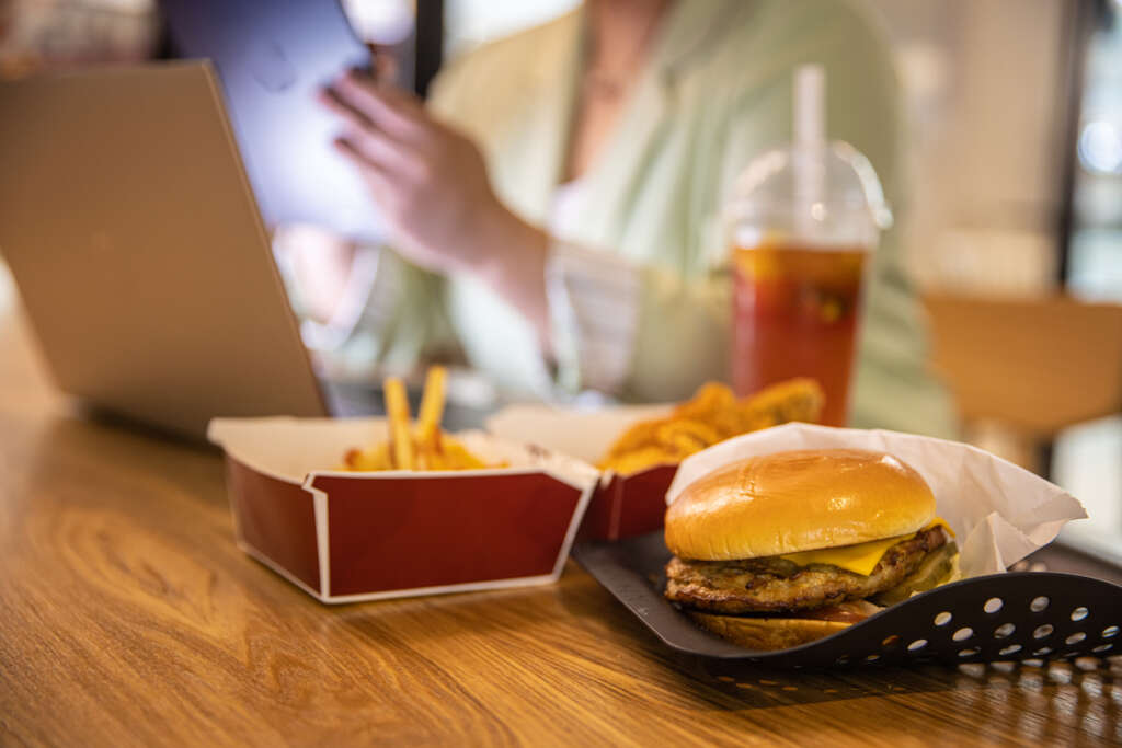 Image of a fast food meal with fries and a cheeseburger placed on a table next to someone using their computer. 