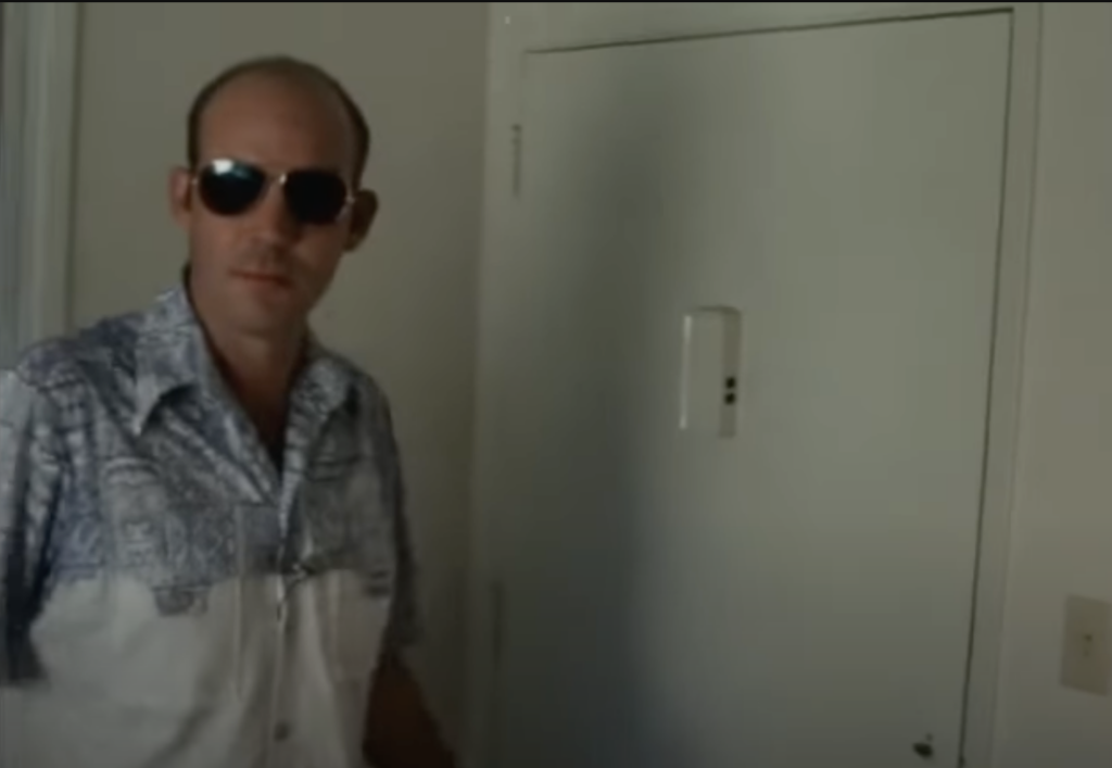 A young Hunter S. Thompson wearing sunglasses inside a hotel room