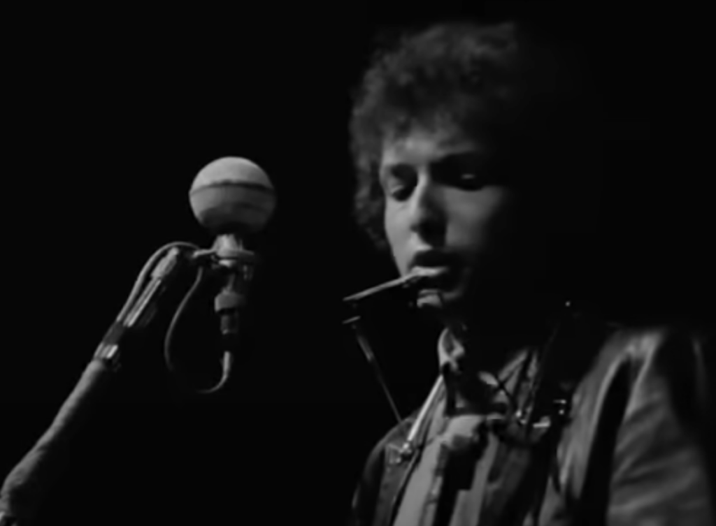 A still black and white image of Bob Dylan in concert. 