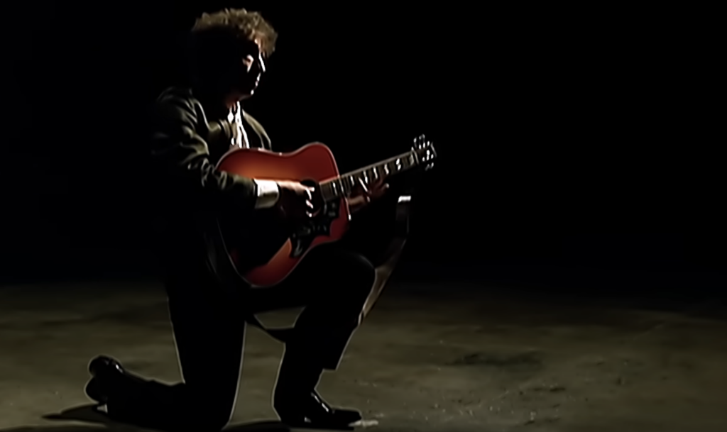 An image of Bob Dylan with his guitar on bended knee. 