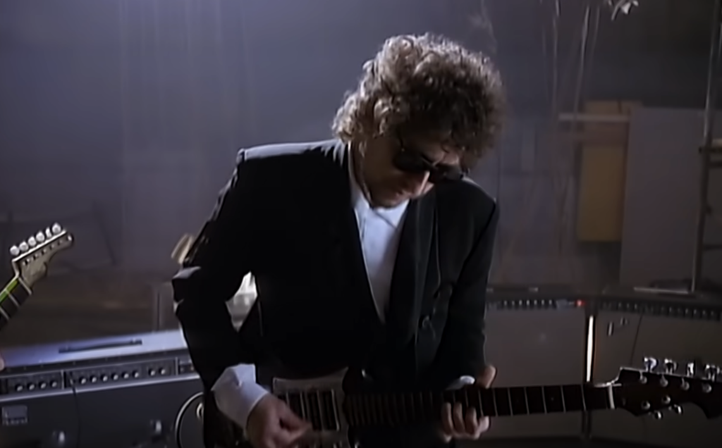 A still image of Bob Dylan playing guitar with his sunglasses on. 