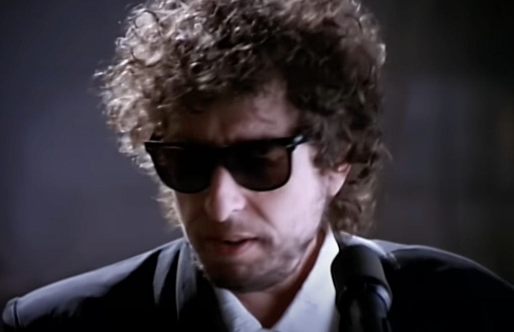 An image of Bob Dylan singing with sunglasses on. 