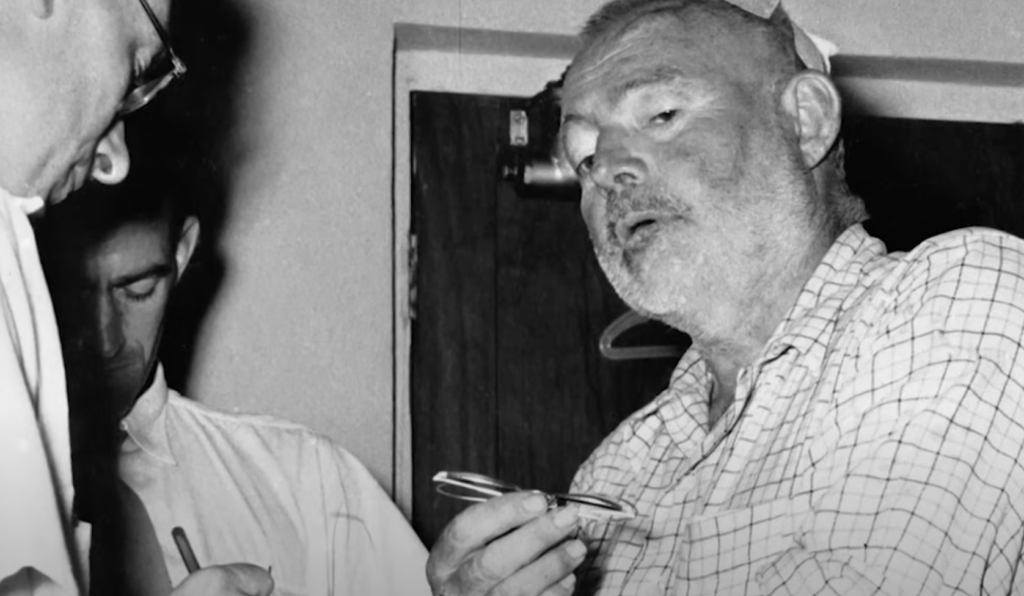 Ernest Hemingway holds glasses and looks into the distance. 