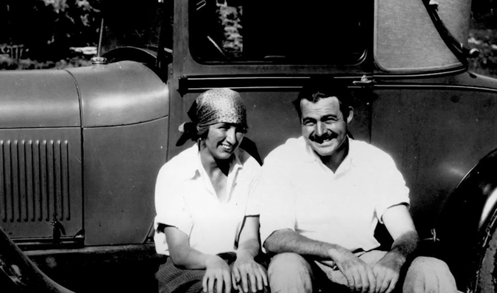 Ernest Hemingway smiling, sitting in front of a car next to a young woman. 