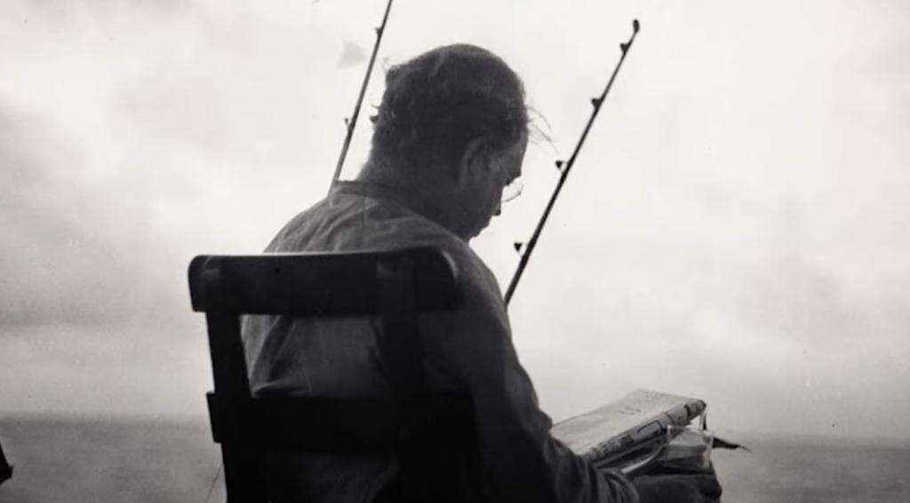 An older Ernest Hemingway sits on chair, reading paper, with fishing rod in front of him. 