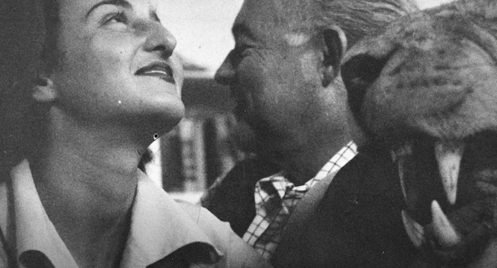 Ernest Hemingway smiles while looking at a woman next to him. 