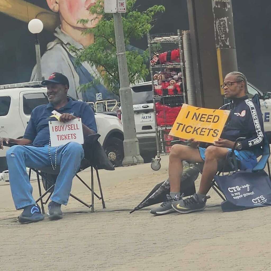 Two men sitting outside a sporting event. One holding a sign that says 'I buy/sell tickets' and the other says 'I need tickets' 