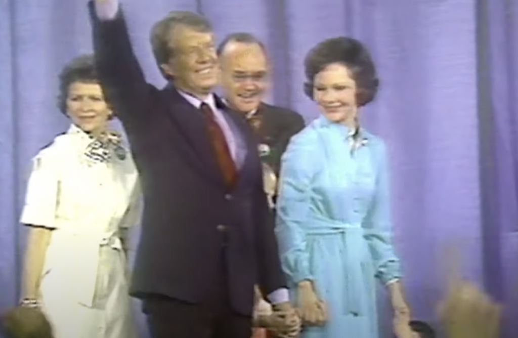 An image of Jimmy Carter walking across the stage with his wife. 