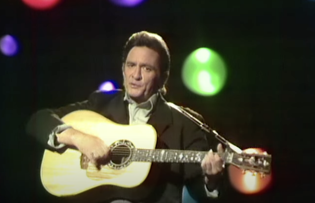 Johnny Cash holding the guitar and singing in front of multi-colored lights. 