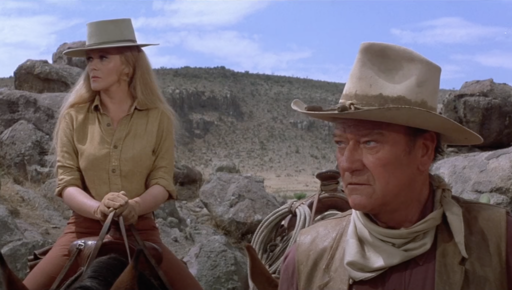 A still image of John Wayne looking off into distance next to a woman on a horse. 