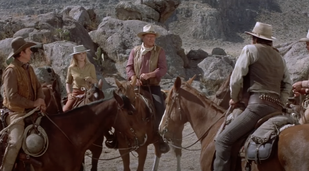 A still image of John Wayne talking to his crew of cowboys. 