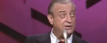 A man with gray hair slicked back is speaking into a microphone. He is wearing a dark suit jacket, a white shirt, and a maroon tie. The background is pink with black geometric shapes. The man appears to be on stage.