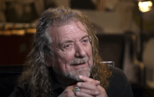 Robert Plant with long, wavy gray hair and a beard is sitting with his hands clasped under his chin. He is smiling and looking slightly to the side. He is wearing a dark-colored shirt and a turquoise ring on his right hand. The background is blurred.