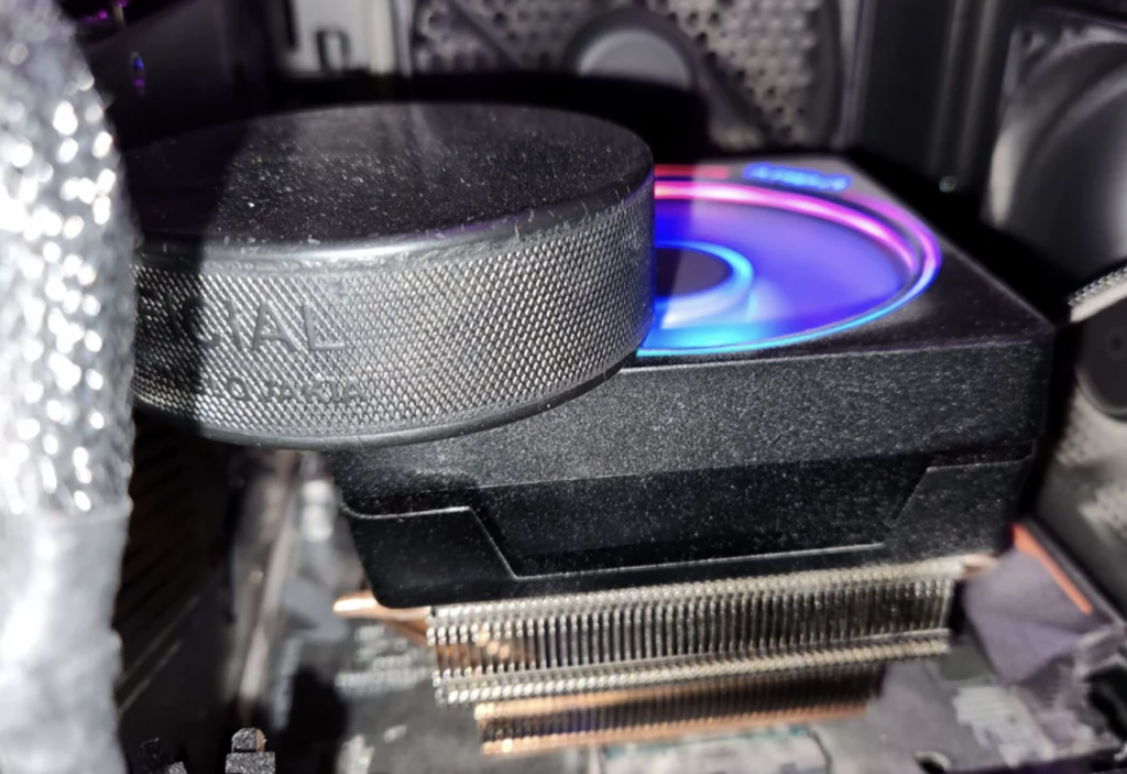 A close-up view inside a computer case showing a CPU cooler with a circular fan emitting rainbow LED lights, mounted on a black heat sink with visible copper heat pipes. The background shows parts of the motherboard and other components.