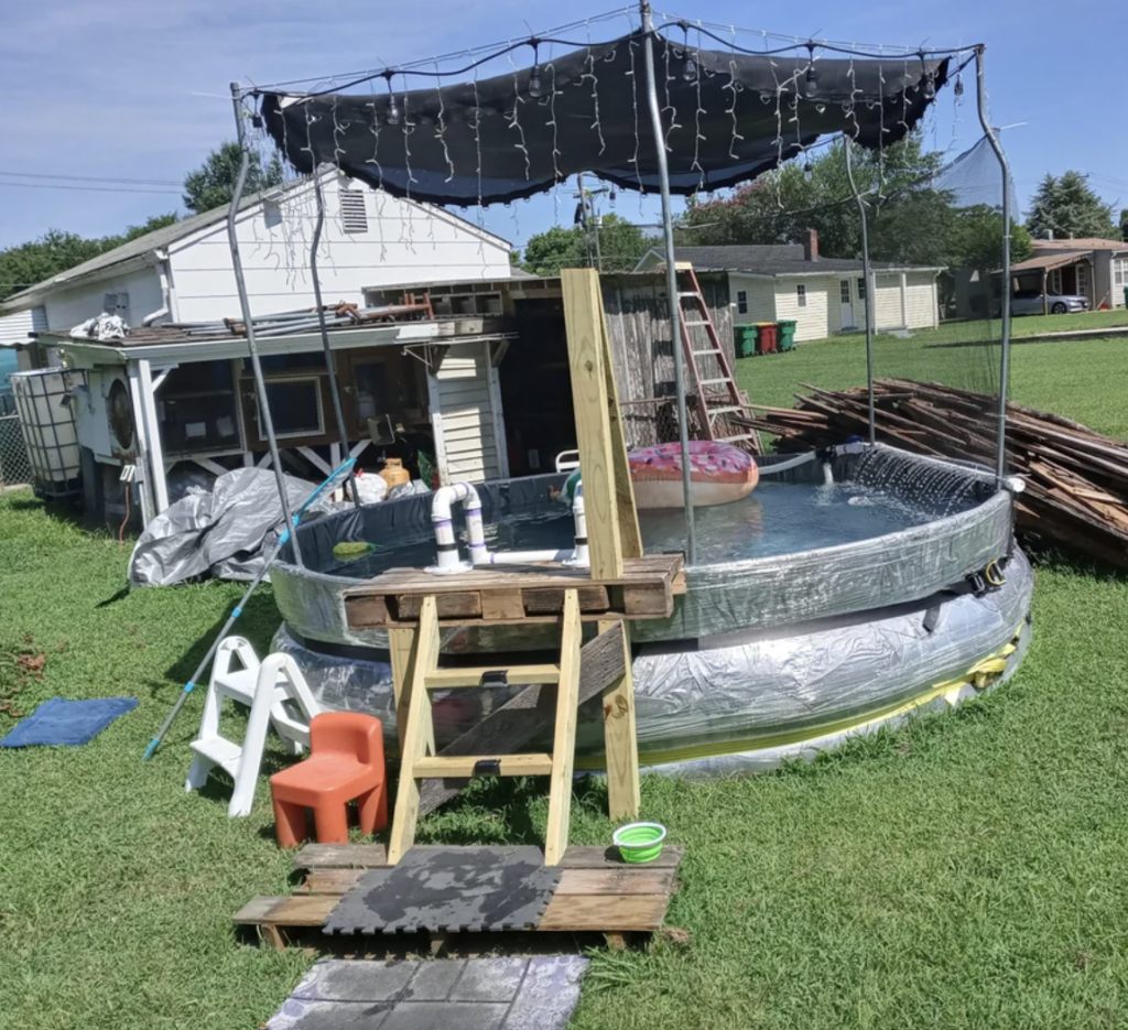 A backyard DIY above-ground pool with a wooden ladder, sunshade canopy, and string lights. There are a small blue and orange step stool, a white plastic chair, and a hose nearby. The area appears to be surrounded by grass and neighboring houses are visible.
