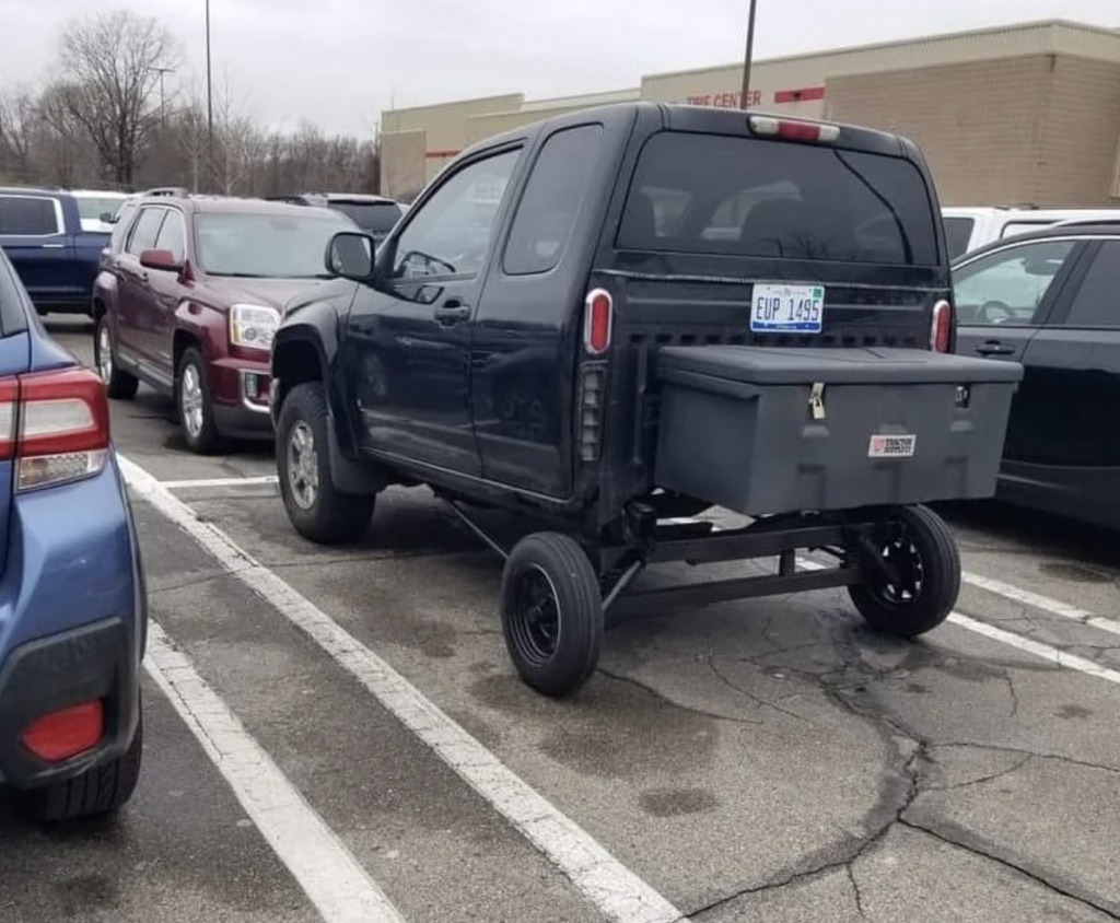 A black pickup truck in a parking lot has been humorously modified with an additional small axle and wheels near the back, giving it a peculiar and unconventional appearance. Other cars are parked nearby, and the environment appears to be a typical commercial area.