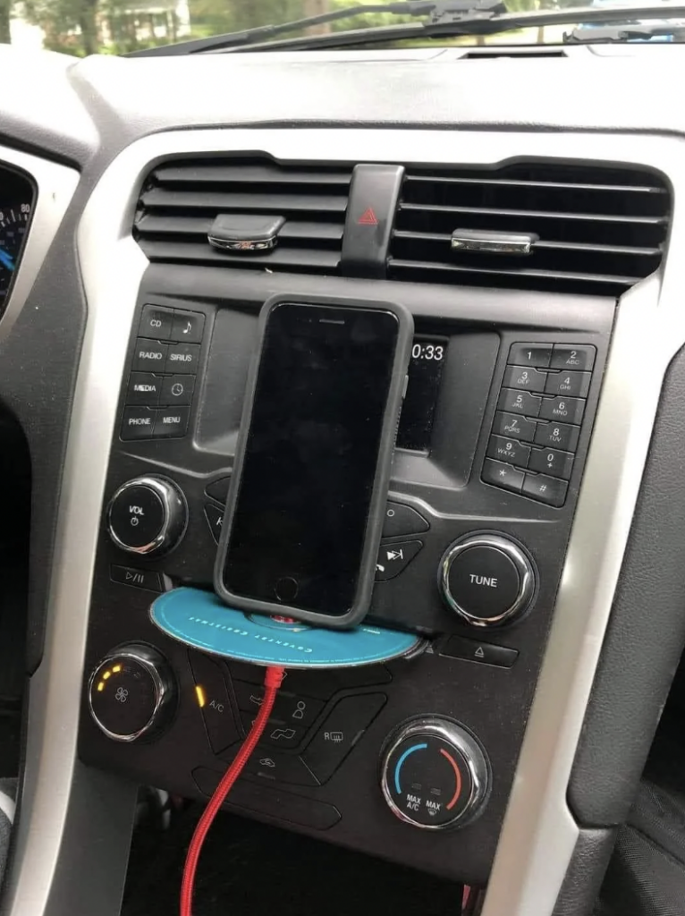 A smartphone mounted on a dashboard holder within a car. The phone is connected to a red charging cable. The car's dashboard features buttons and knobs for radio, media, and climate controls, with visible air vents above. The background shows a glimpse of trees through the windshield.