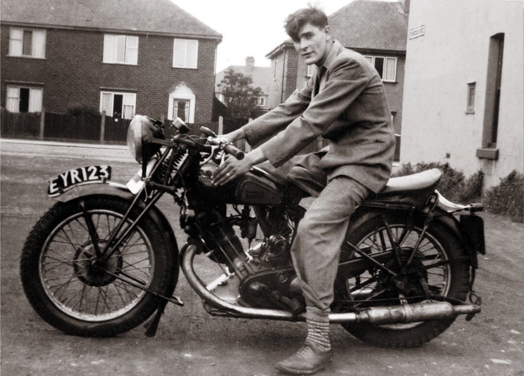 A person wearing a jacket and pants is sitting on a vintage motorcycle with the license plate EYR 123. The background shows houses with brick walls and fenced yards on a quiet street. The image has a black and white, nostalgic appearance.