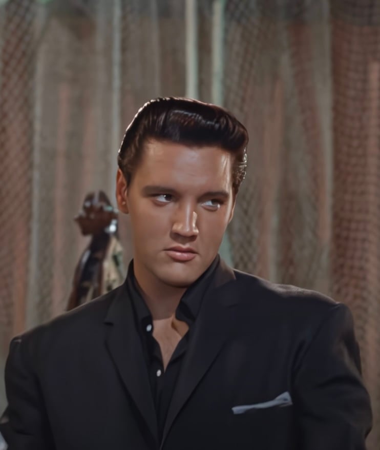 A person with slicked-back dark hair wearing a black suit and a dark shirt looks to the side with a serious expression. The background is a textured curtain and a silhouette of another figure.