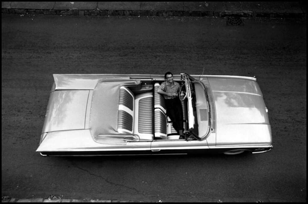 Aerial view of a vintage convertible car with the top down. A man in a plaid shirt is seated in the driver's seat, looking up at the camera. The car has wide, striped seats and is parked on a street.