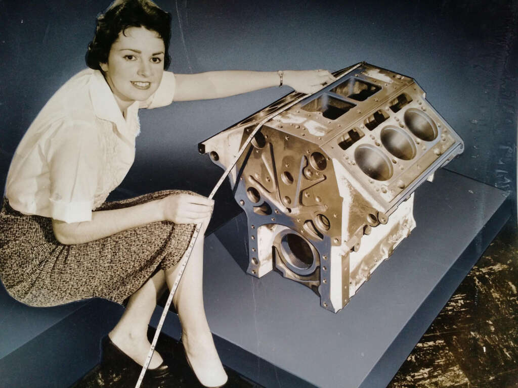 A woman in a white blouse and patterned skirt kneels beside a large metallic engine block, measuring it with a tape. The setting is a studio, with the engine block placed on a raised platform.