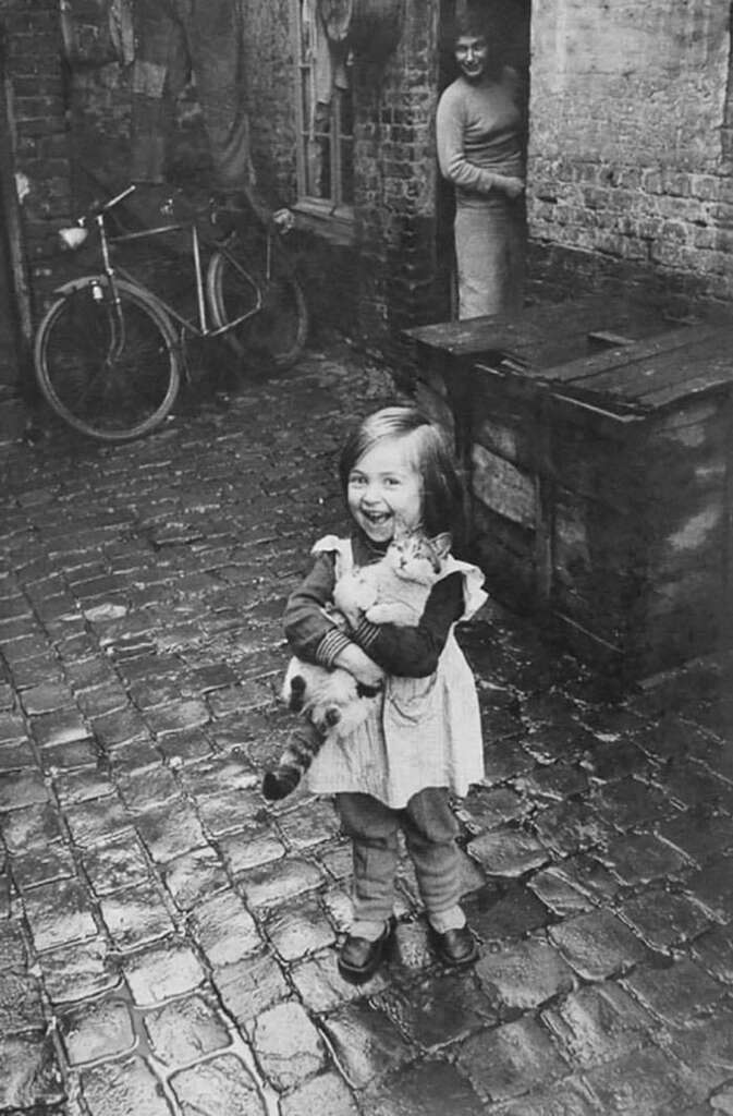 A young girl smiles joyfully while holding a cat in her arms. She stands on a cobblestone street with a bicycle leaning against a brick wall in the background. An adult observes from a doorway nearby, partially visible.