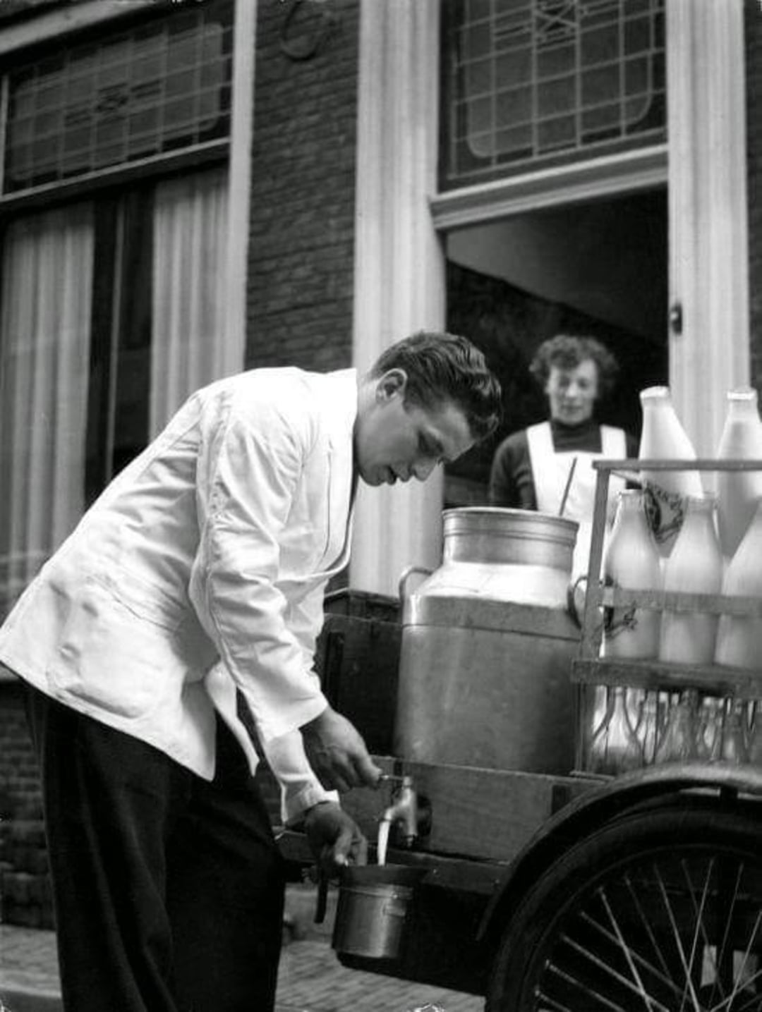 A man in a white coat fills a metal cup from a large milk can on a cart, while a woman stands in a doorway behind him. Several bottles of milk are visible on the cart. The scene appears to be from the past.