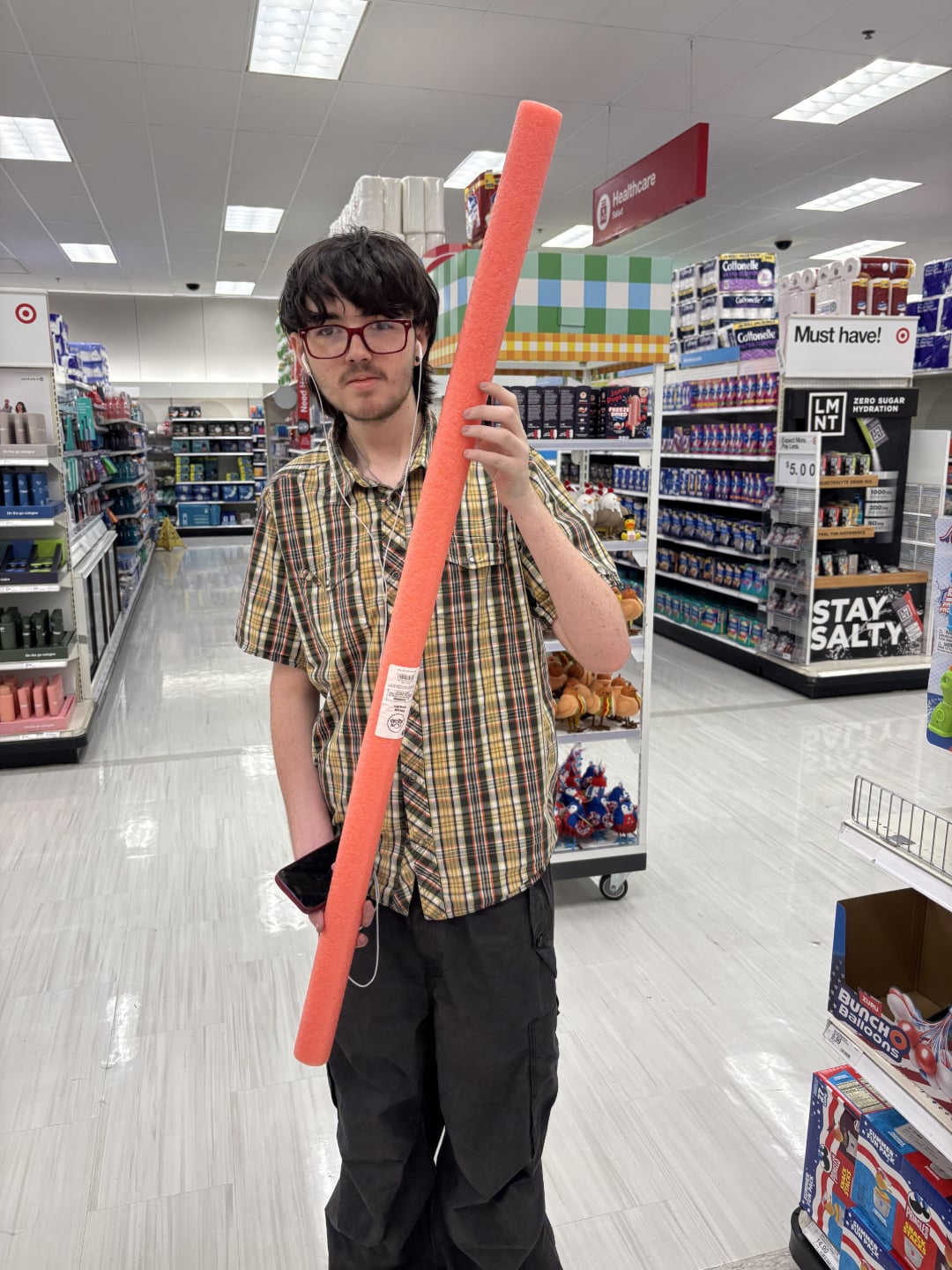 A person wearing glasses and a plaid shirt stands in a store aisle holding a long orange pool noodle, with various products on shelves in the background.