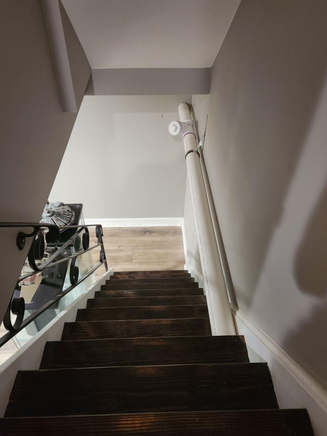 A view looking down a narrow wooden staircase with dark steps, a white handrail on the right, and a light fixture on the wall ahead. The stairs lead to a room with light-colored wood flooring.