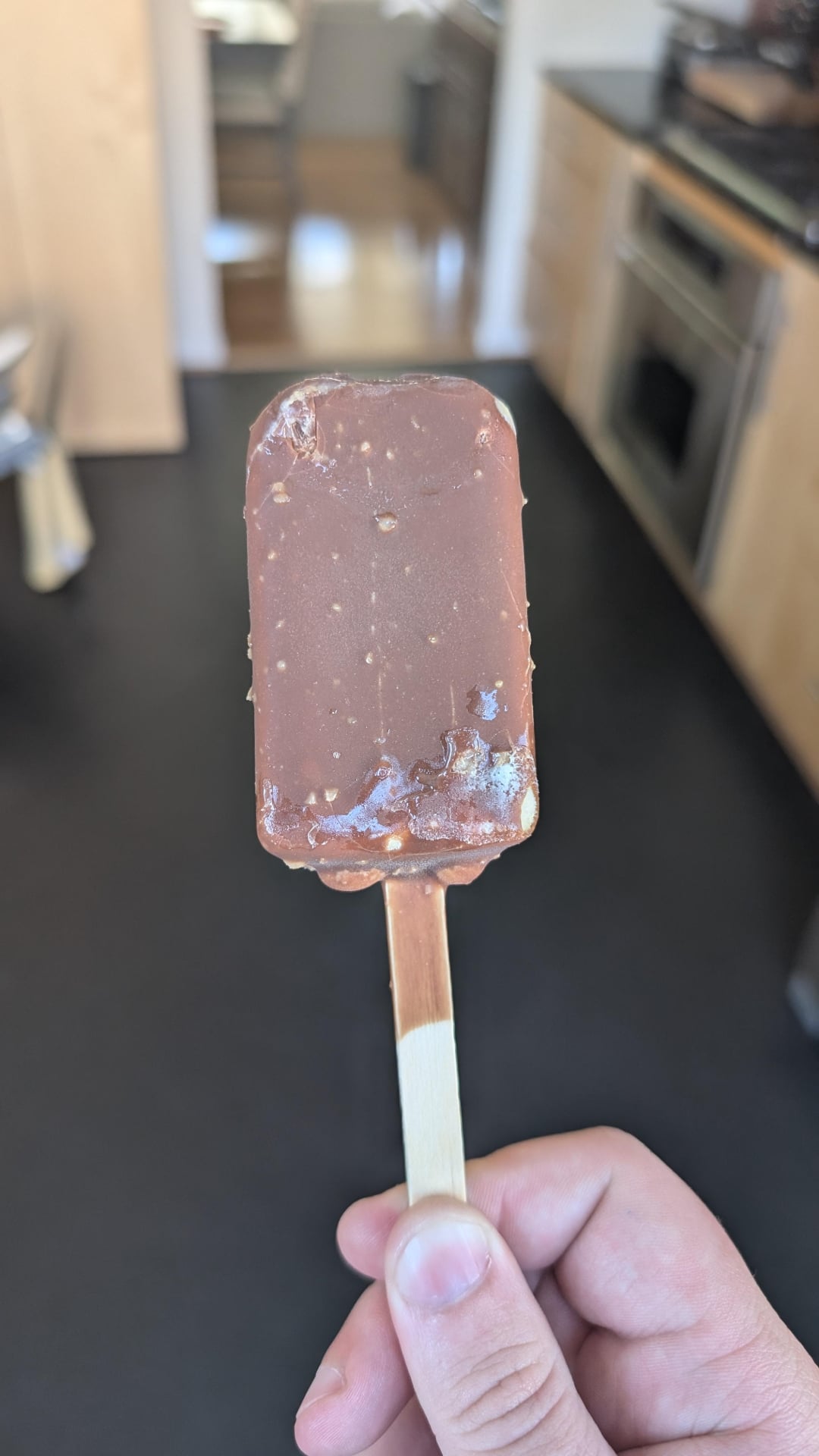 A hand holds a partially melted chocolate-covered ice cream bar on a wooden stick in a modern kitchen with blurred countertops and appliances in the background.