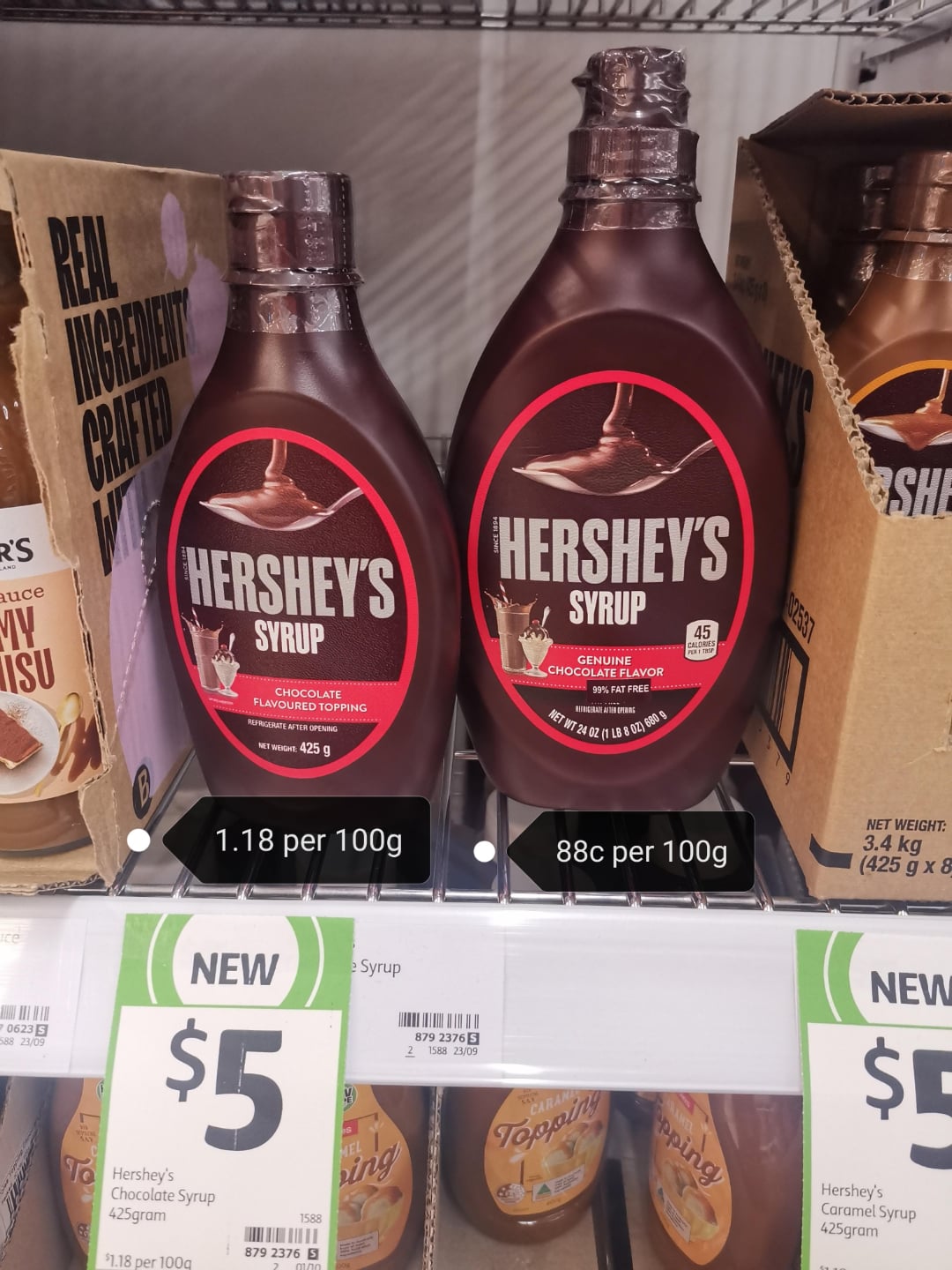 Two bottles of Hershey’s Chocolate Syrup are displayed on a supermarket shelf, each priced at $5. The bottle on the left costs $1.18 per 100g, while the bottle on the right costs 88c per 100g.