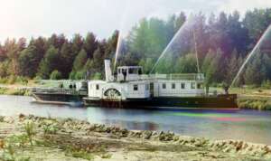 A vintage riverboat travels along a calm river, with trees in the background. The side features a large paddle wheel, and water sprays upward from spigots on the deck. The scene has a colorful, slightly faded quality.