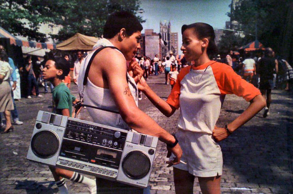 A man wearing a tank top with a large boombox over his shoulder smokes, while a woman in a red and white shirt stands with one hand on her hip talking to him. They are outside on a cobblestone street with people and tents in the background.
