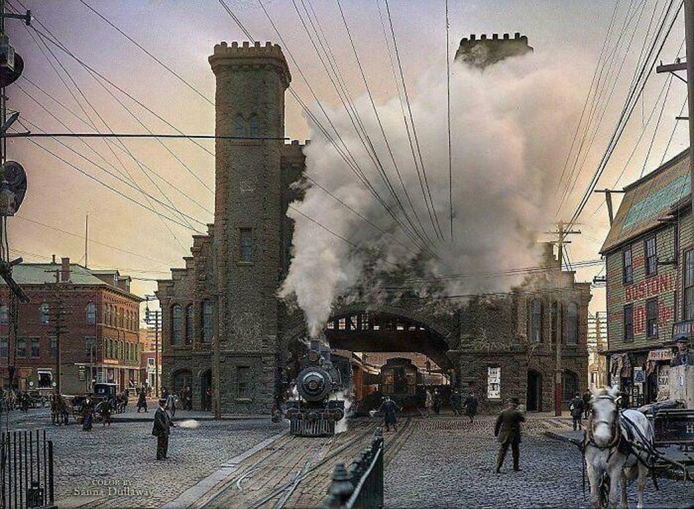 A historical scene featuring a steam locomotive emitting smoke while passing through an archway of a large, castle-like brick building. People and horse-drawn carriages are visible on the cobblestone street lined with vintage buildings.