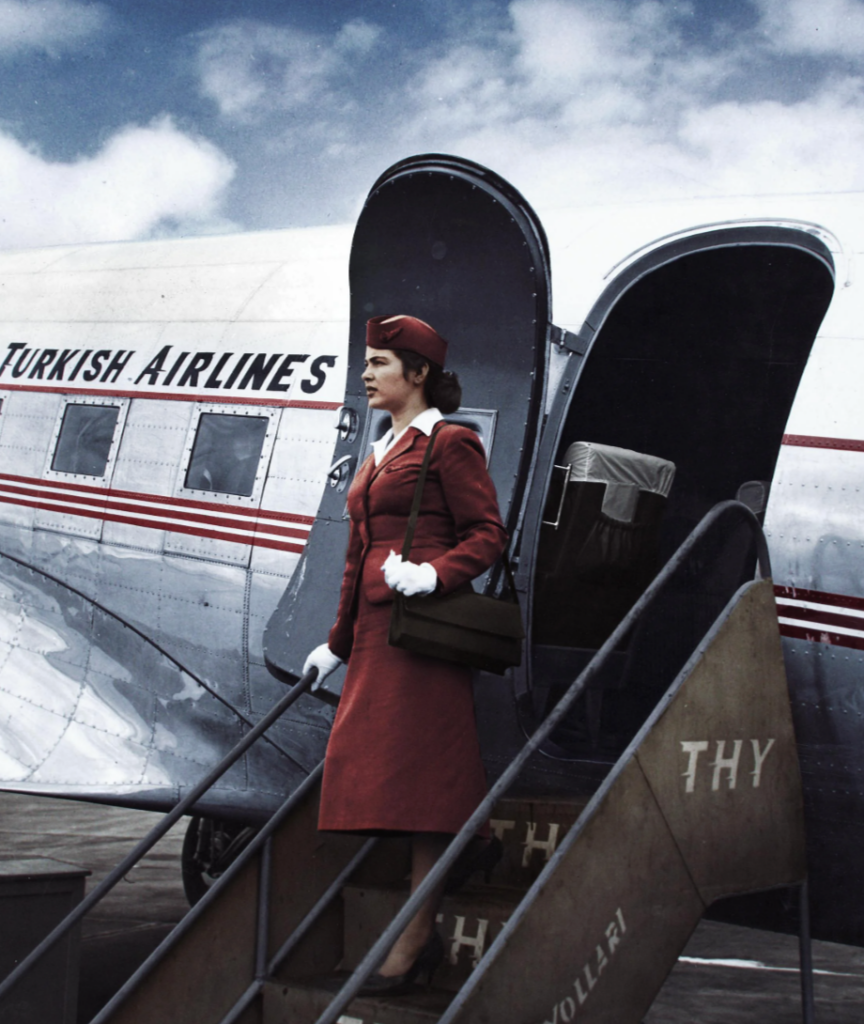 A flight attendant in a red uniform and hat descends the stairs of a Turkish Airlines plane. The aircraft is shiny with visible red stripes and the company's name. The sky is partly cloudy in the background.