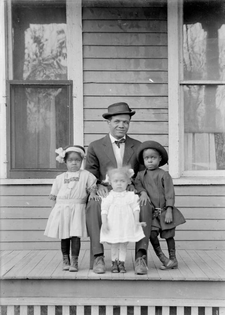 A man wearing a suit and hat sits on a porch with three young children. Two girls stand on either side, wearing dresses and hats. A younger child stands in the center wearing a light dress. The wooden house wall and windows form the background.
