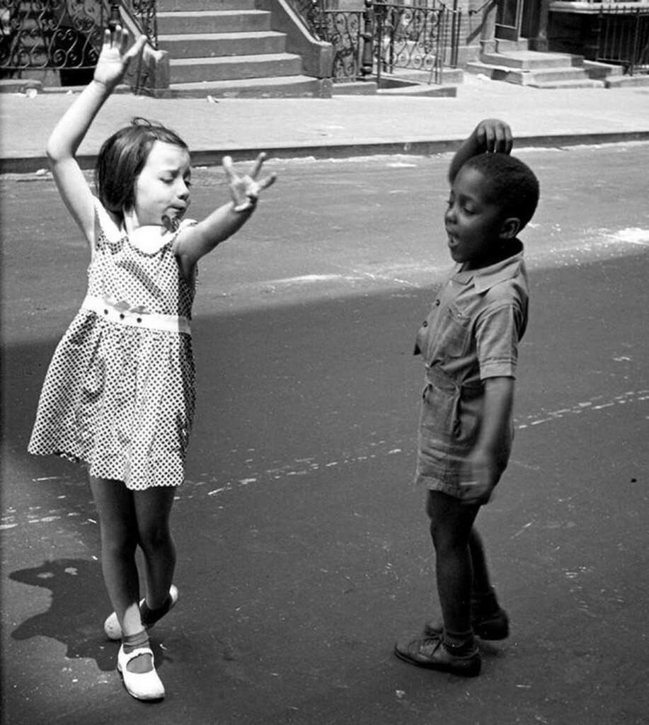 A young girl in a polka dot dress and a boy in a short-sleeved outfit joyfully dance on a street. The background shows stairs and railings of a brownstone-style building.