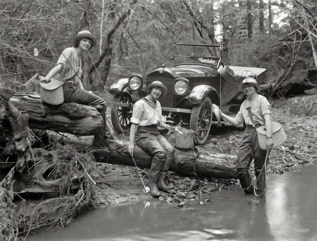 Three women in early 20th-century attire pose with fishing gear on a log over a creek. Behind them, a vintage car is parked in a wooded area. They appear cheerful and are wearing hats and waders.