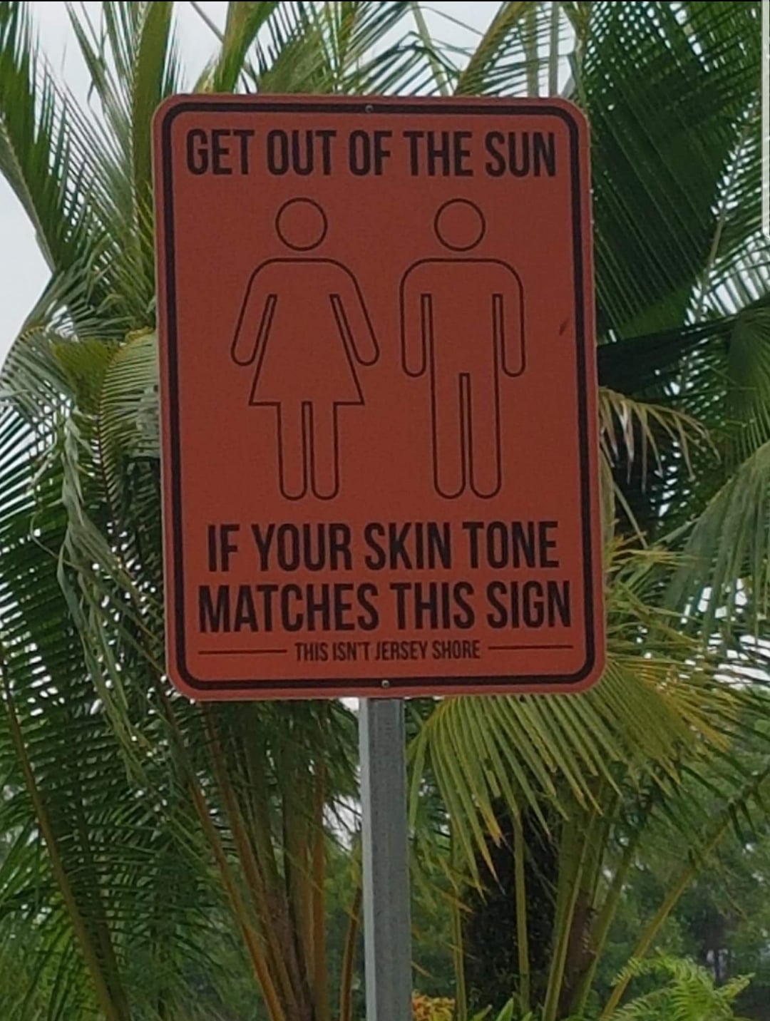 A red sign shows male and female icons and reads: "GET OUT OF THE SUN IF YOUR SKIN TONE MATCHES THIS SIGN. THIS ISN'T JERSEY SHORE." Palm trees are visible in the background.