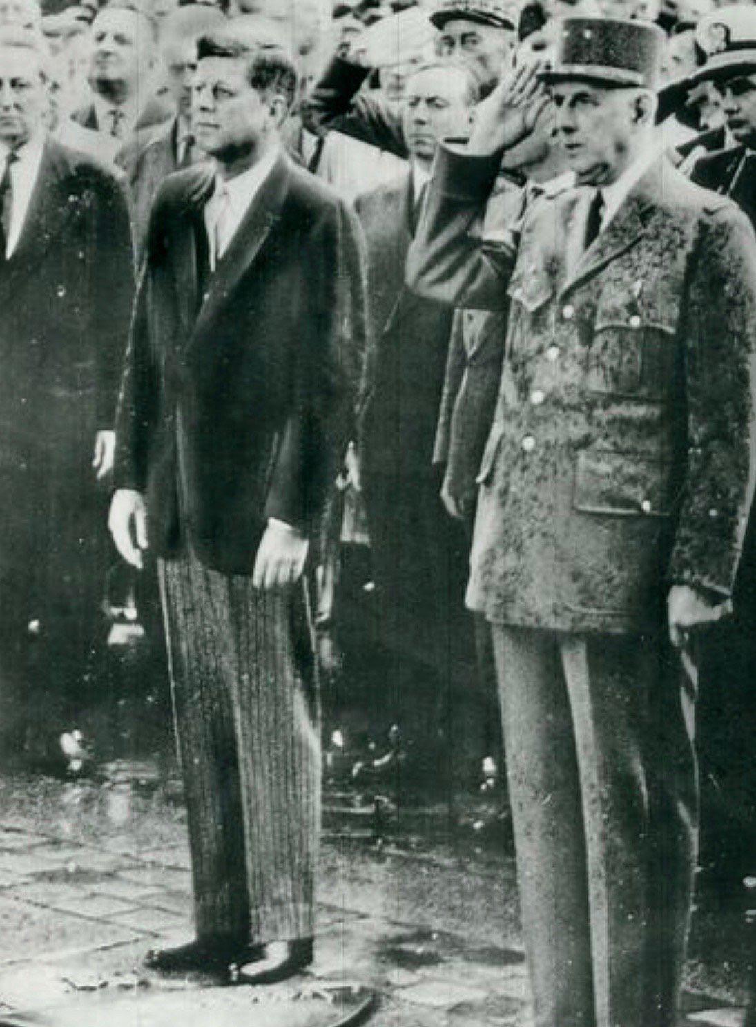 Two men stand side by side in a formal setting. One wears a suit, the other a decorated military uniform and cap, saluting. A crowd of men in suits stands behind them, watching the ceremony. The image is black and white.