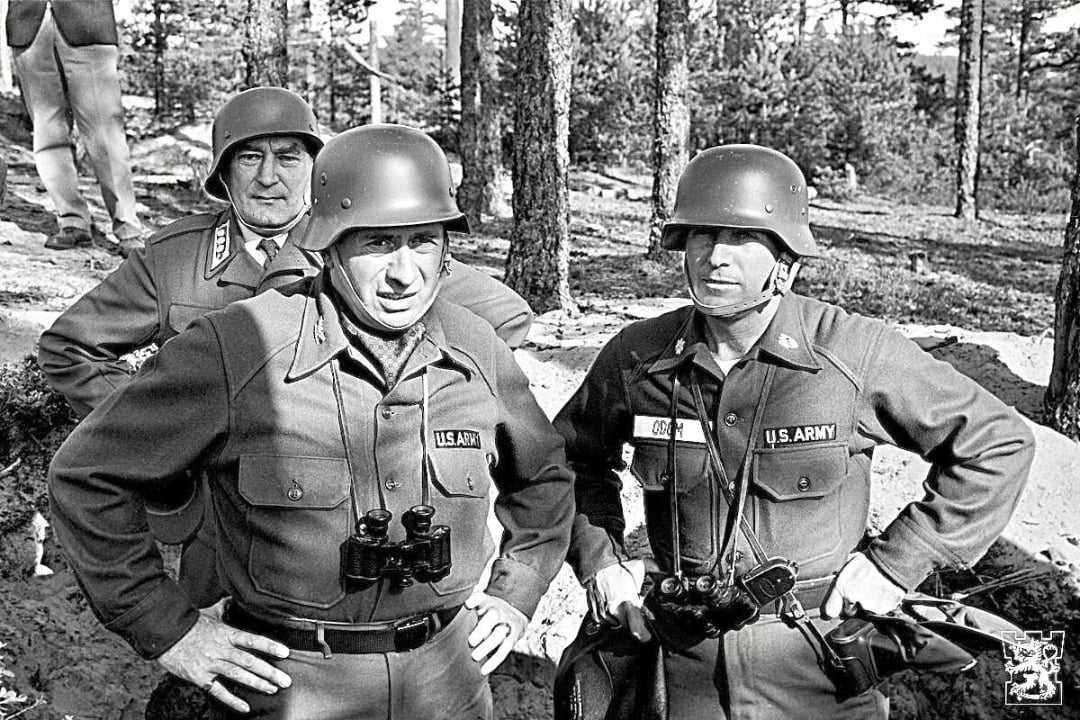 Three U.S. Army soldiers in uniform and helmets stand in a wooded outdoor area, two with binoculars around their necks, looking focused and serious; trees and sunlight are visible in the background.