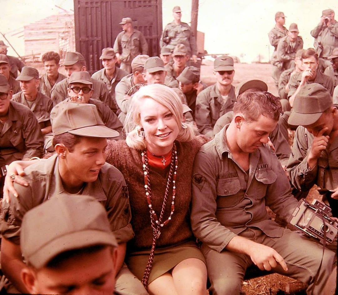 A woman with blonde hair sits smiling among a group of uniformed soldiers, some looking at the camera and others looking elsewhere, in an outdoor setting with buildings and equipment in the background.