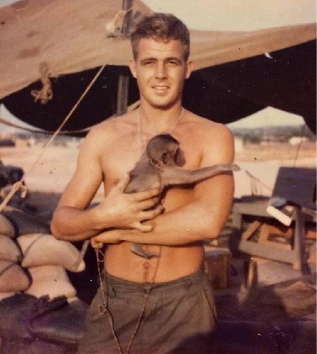 A shirtless young man stands outdoors, smiling and holding a small monkey in his arms. Behind him are military-style tents and equipment, suggesting a camp setting.