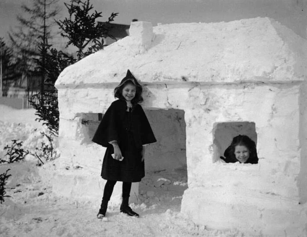 Two children play with a snow house in winter. One stands outside wearing a dark coat and hat, and the other peeks through the window, smiling. Snow covers the ground and a pine tree is nearby.