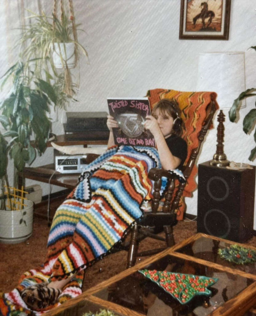 A person with headphones sits in a retro-style living room, covered with a colorful crochet blanket, holding a "Twisted Sister" album. There are houseplants, a speaker, and a large table with several bowls in the room.