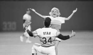 A person in a baseball uniform with "RYAN 34" on the back kneels, arms open, as a woman with light-colored hair runs toward them on the baseball field, also with arms open. The background shows other players and part of the stadium.