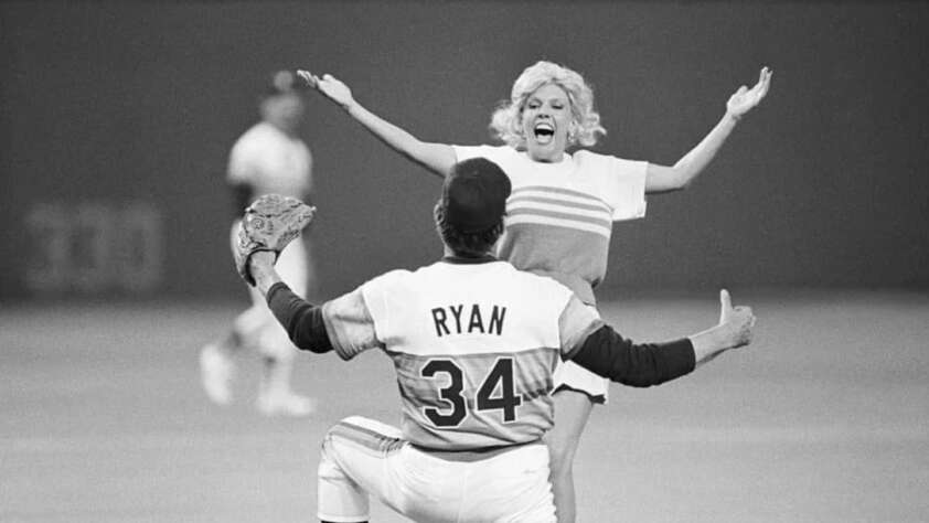 A person in a baseball uniform with "RYAN 34" on the back kneels, arms open, as a woman with light-colored hair runs toward them on the baseball field, also with arms open. The background shows other players and part of the stadium.