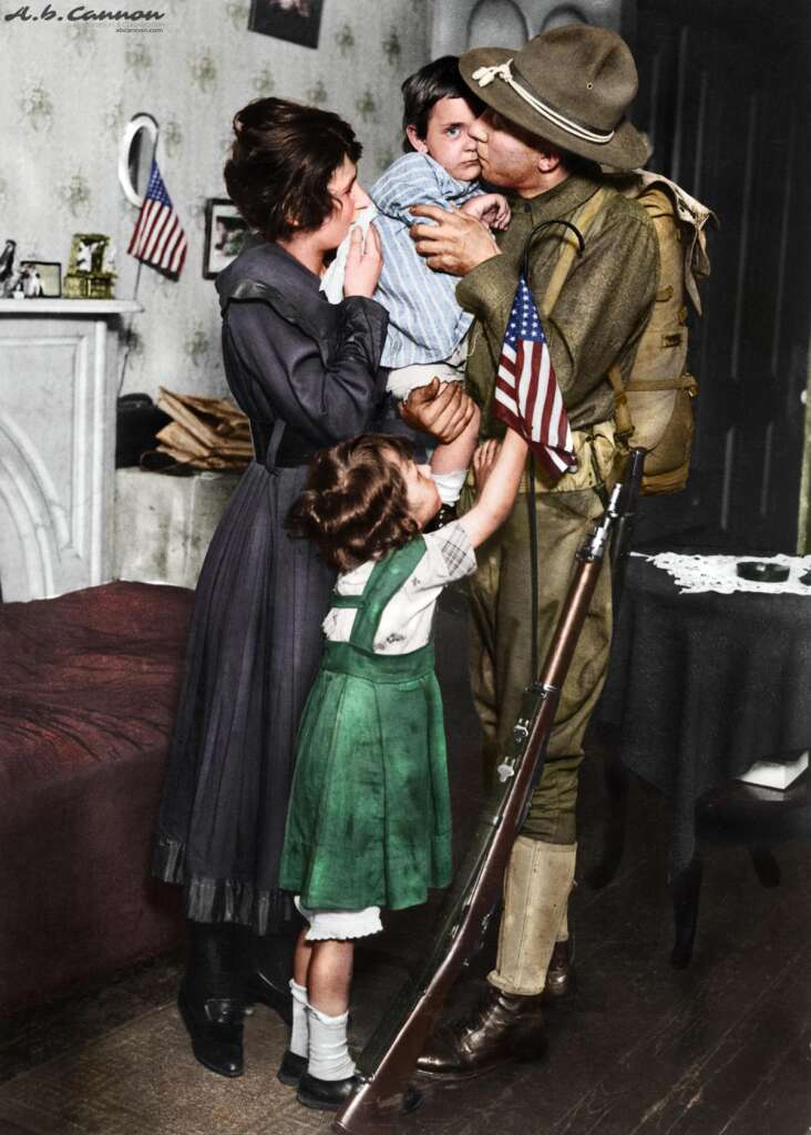 A soldier in uniform holds a young child as a woman and a girl reach towards them in a room decorated with American flags. A rifle is propped nearby. The woman appears emotional, and the girl is dressed in a green dress.
