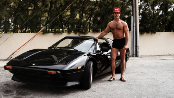80s, cars, bkies, vintage, nostalgia, 1980s. A muscular man in a red cap, black shorts, and flip-flops stands next to a black sports car, outdoors, with trees and a beige wall in the background.