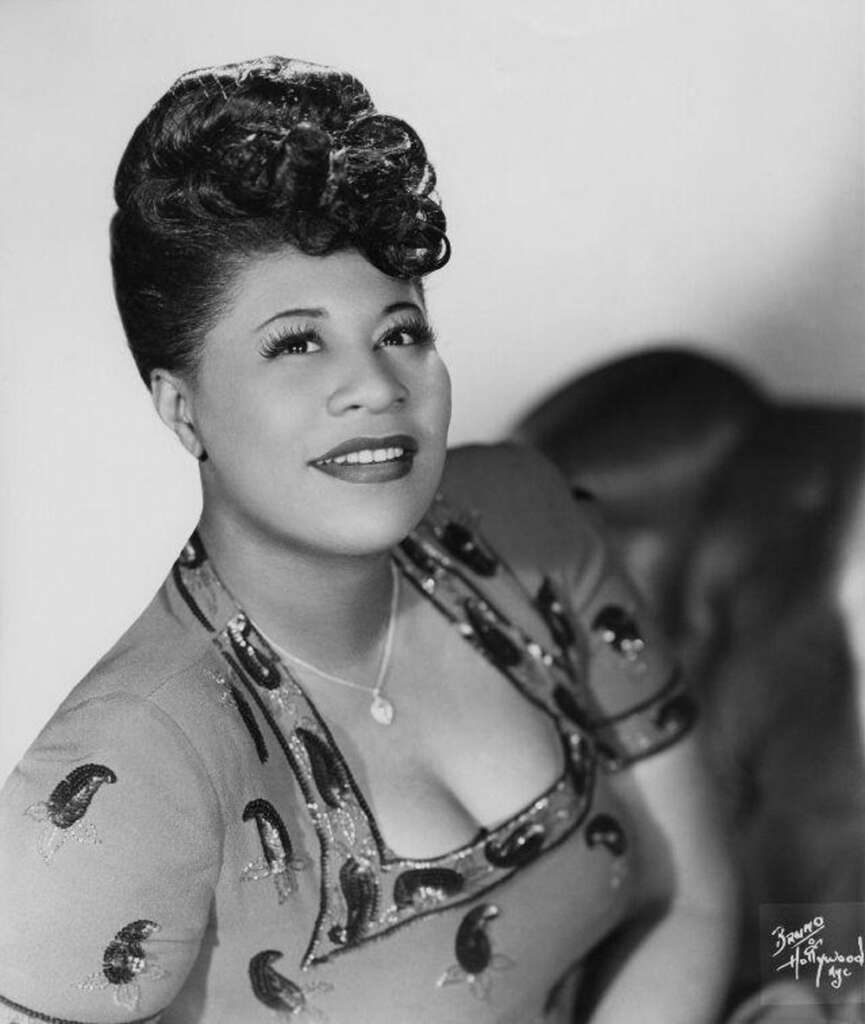 Black and white portrait of a woman smiling upwards. Her hair is styled in an elegant updo. She wears a patterned dress with leaf motifs and a necklace. The background is blurred.